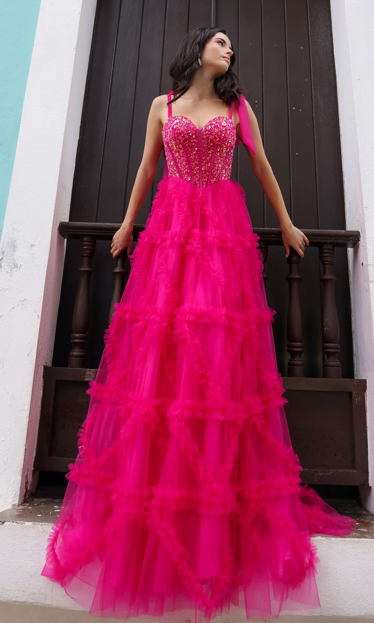 A woman in the Shoulder-Bows Long Ruffled Prom Gown T1340, a bright pink floor-length tulle dress with sequin details and a beaded corset bodice, stands before a dark wooden door, looking to her right.